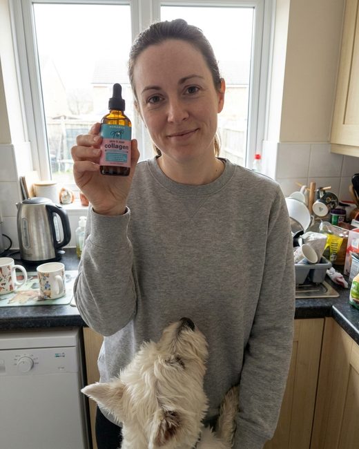 A woman in a grey sweatshirt holding the Noramyx bottle in her kitchen, a West Highland White Terrier at her side.