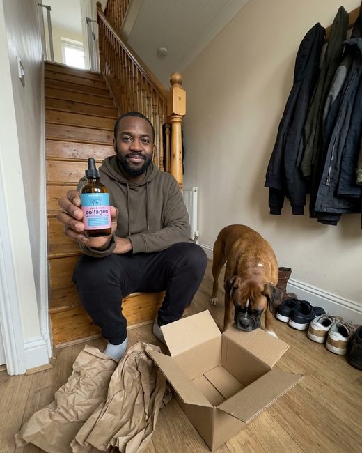 A man sitting on the stairs holding the Noramyx bottle, his Boxer sniffing the opened delivery box.