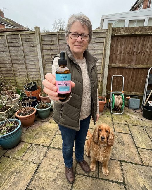 A woman in a gilet holding the Noramyx bottle on her patio, a Cocker Spaniel sitting beside her.