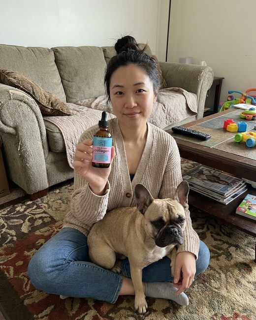A woman sitting on a living-room floor holding the Noramyx bottle, a French Bulldog on her lap.